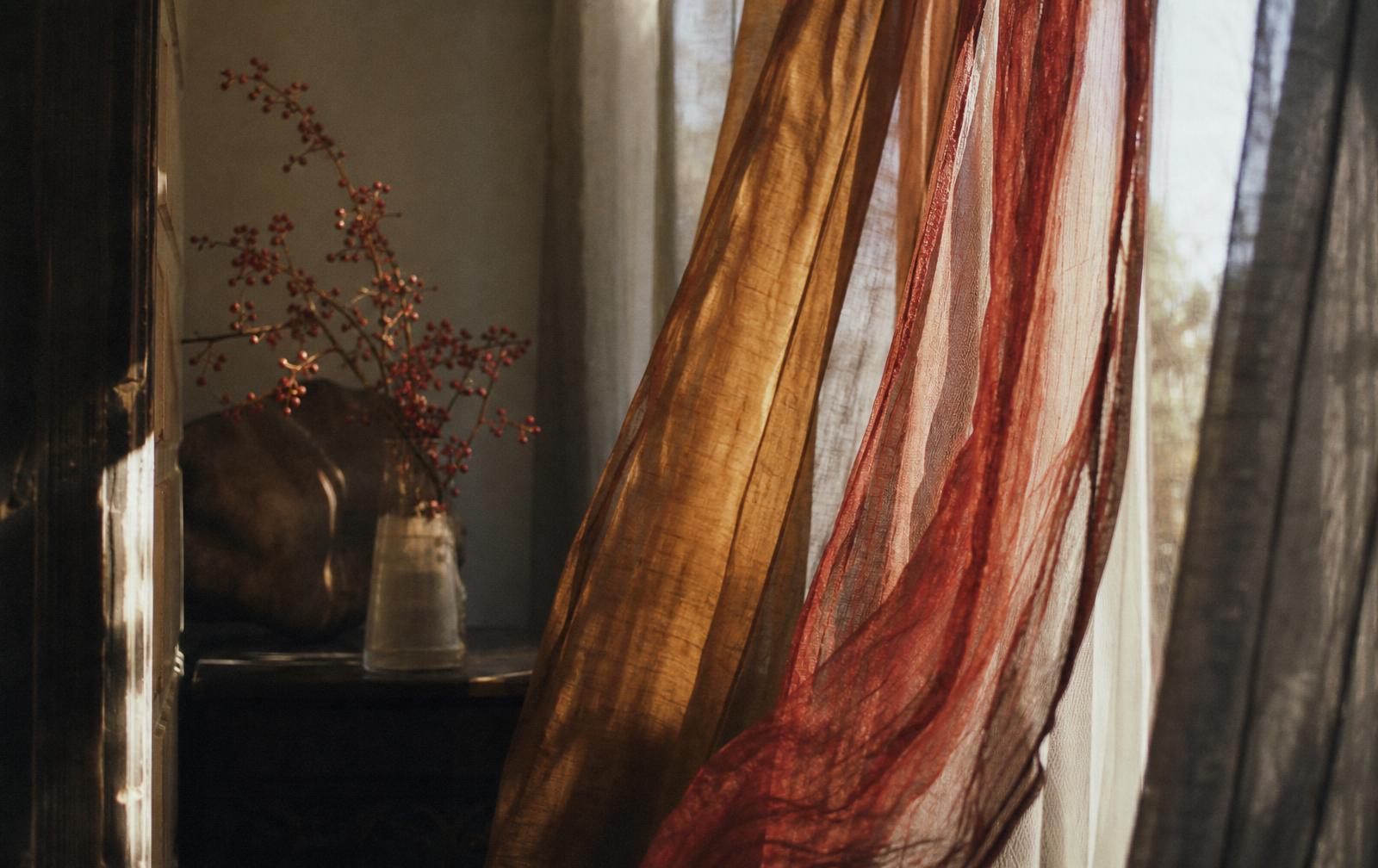 Sheer amber and crimson curtains drifting by a windowsill with a small vase of red berries