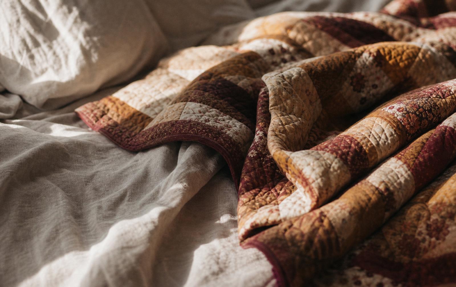 A folded patchwork quilt on unmade linen bedding in soft morning light